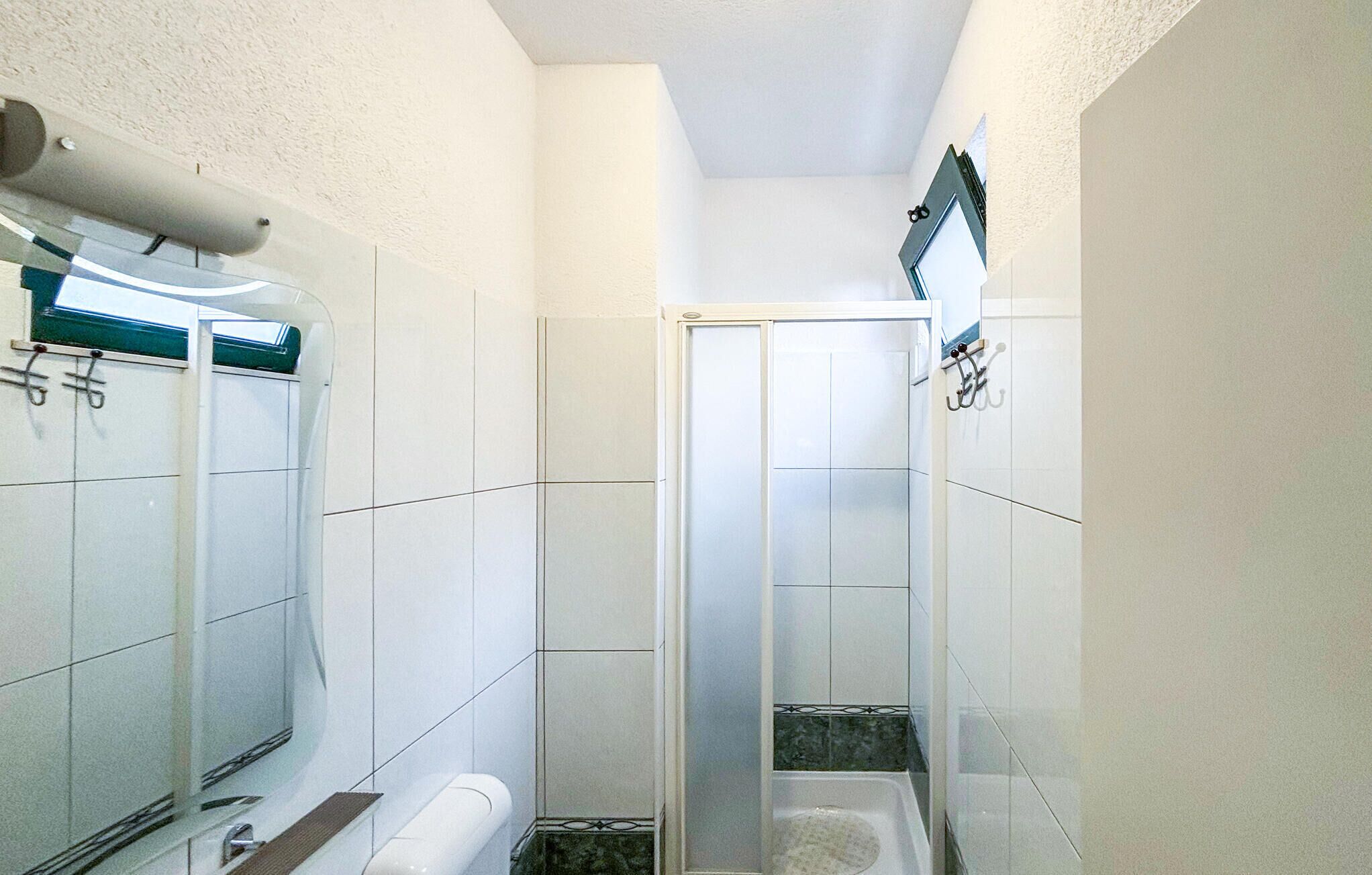 Une petite salle de bain étroite aux murs recouverts de carreaux blancs, dotée d’une douche en verre et de toilettes.