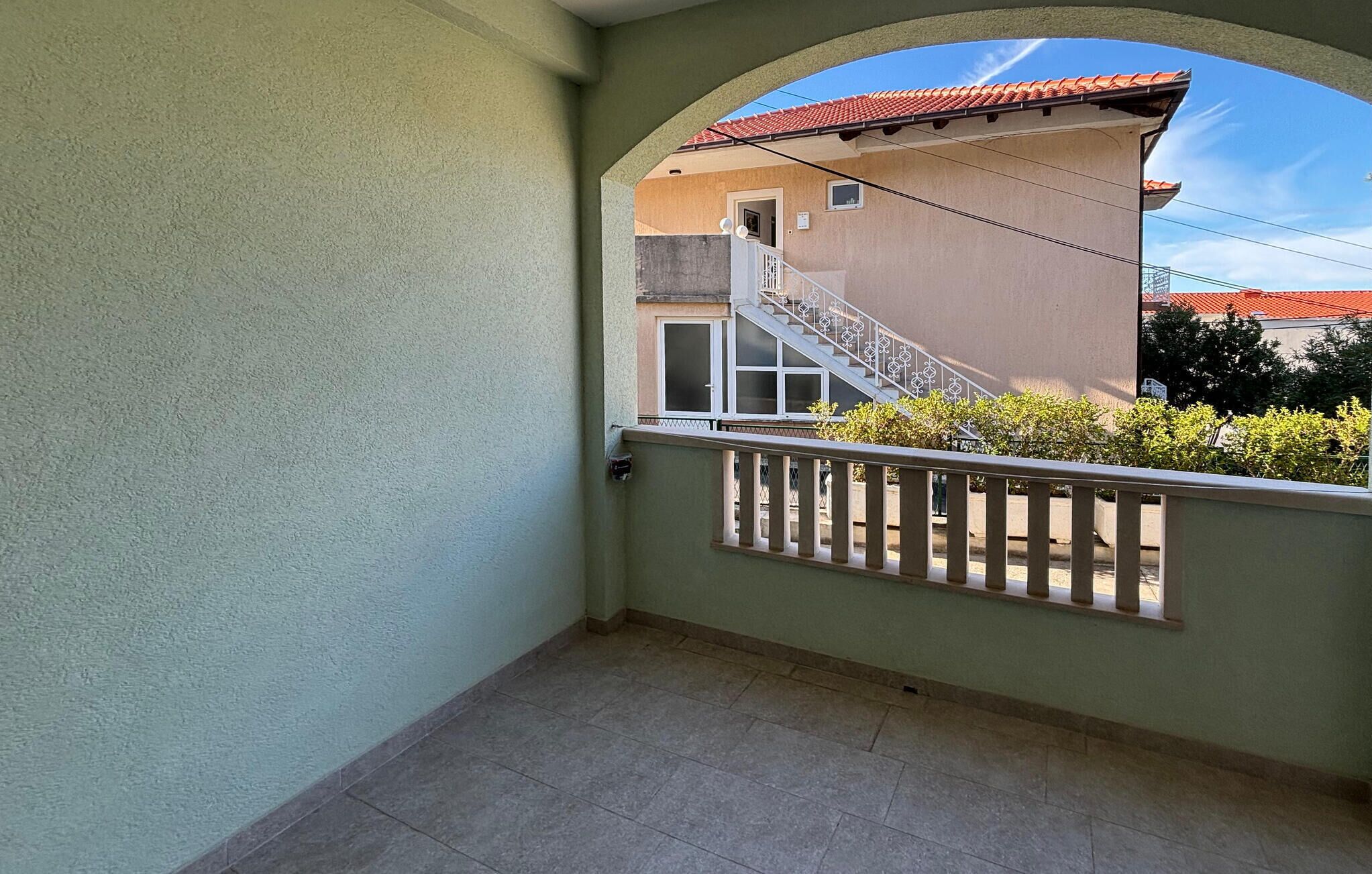 Ein Balkon mit Fliesenboden, Metallgeländer und Blick auf ein Gebäude mit einem roten Ziegeldach.