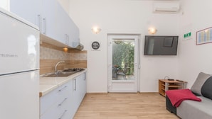 Cocina compacta con estufa integrada, fregadero y refrigerador. Se ve una puerta que da a un espacio exterior con vistas a vegetación. Un televisor de pantalla plana está montado en la pared, y se distingue una pequeña mesa de madera con una manta roja.