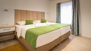 Une chambre d’hôtel moderne avec une tête de lit en bois, un grand lit avec une literie verte et blanche, une table de chevet avec une lampe, et une vue sur l’extérieur par une fenêtre avec des rideaux.