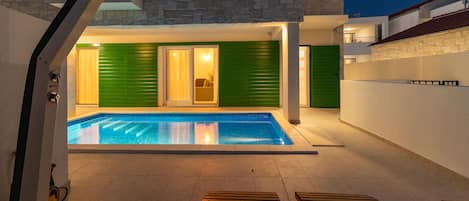 A modern outdoor pool area with a stone wall, green shutters, and a wooden deck.