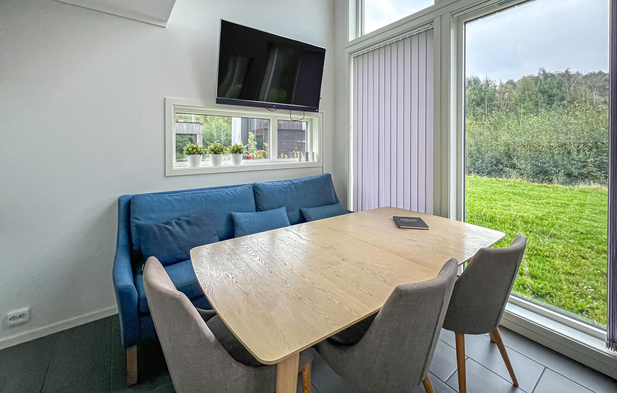 A room with a wooden table, grey chairs, a blue sofa, a TV mounted on the wall, and a window with a view of greenery.