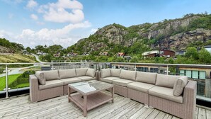 Een balkon met een rieten bank en een bijzettafeltje, met uitzicht op een bergachtig landschap met huizen.