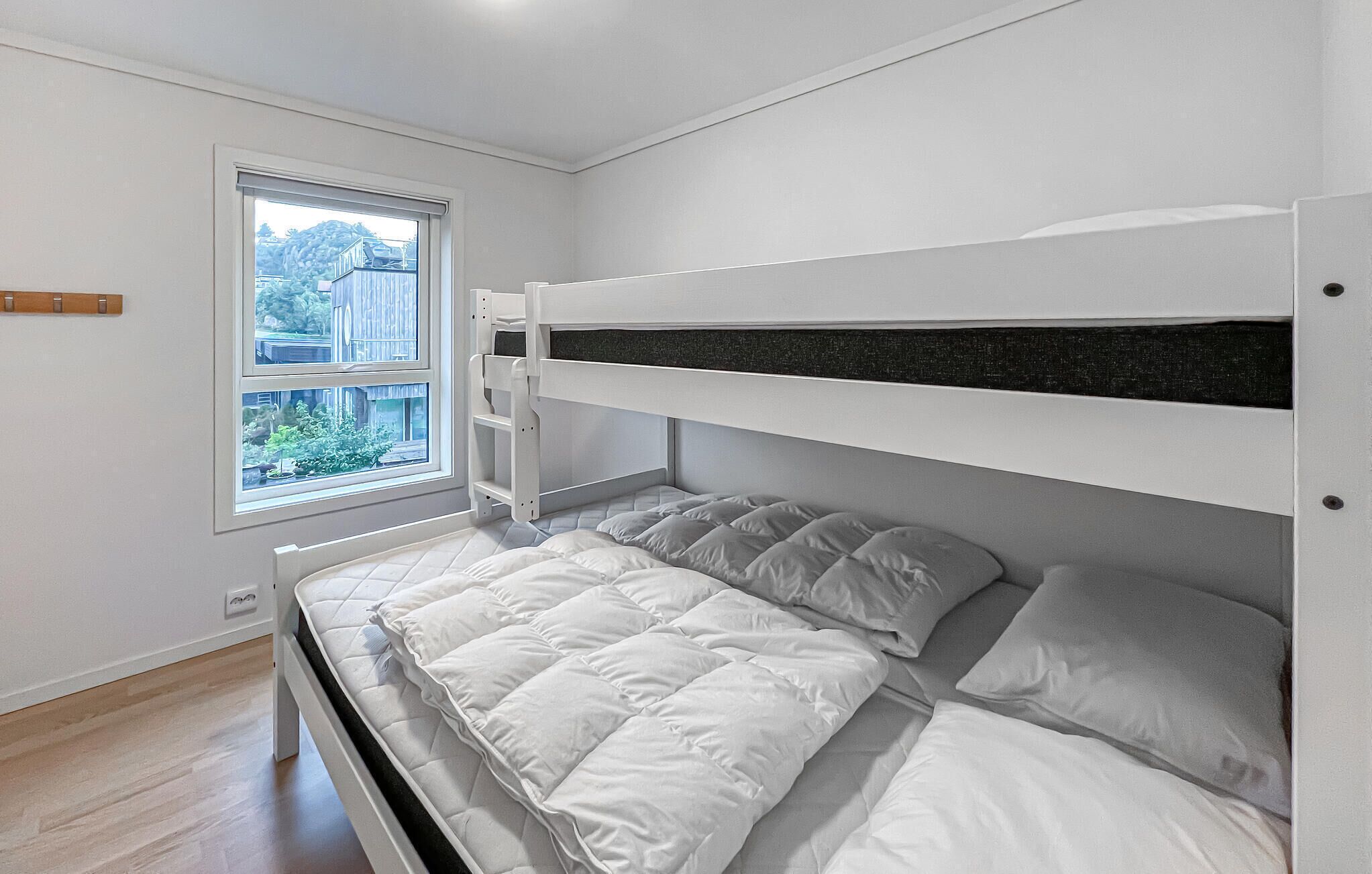 A bunk bed with white bedding and a window with a view of buildings and greenery.