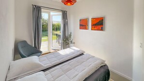 Un dormitorio con una cama, una silla, un ventanal con cortinas y dos cuadros en la pared.