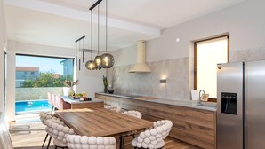 Ett modernt kök med ett matbord i trä, vita sittpallar, ett kylskåp i rostfritt stål och utsikt över en swimmingpool.