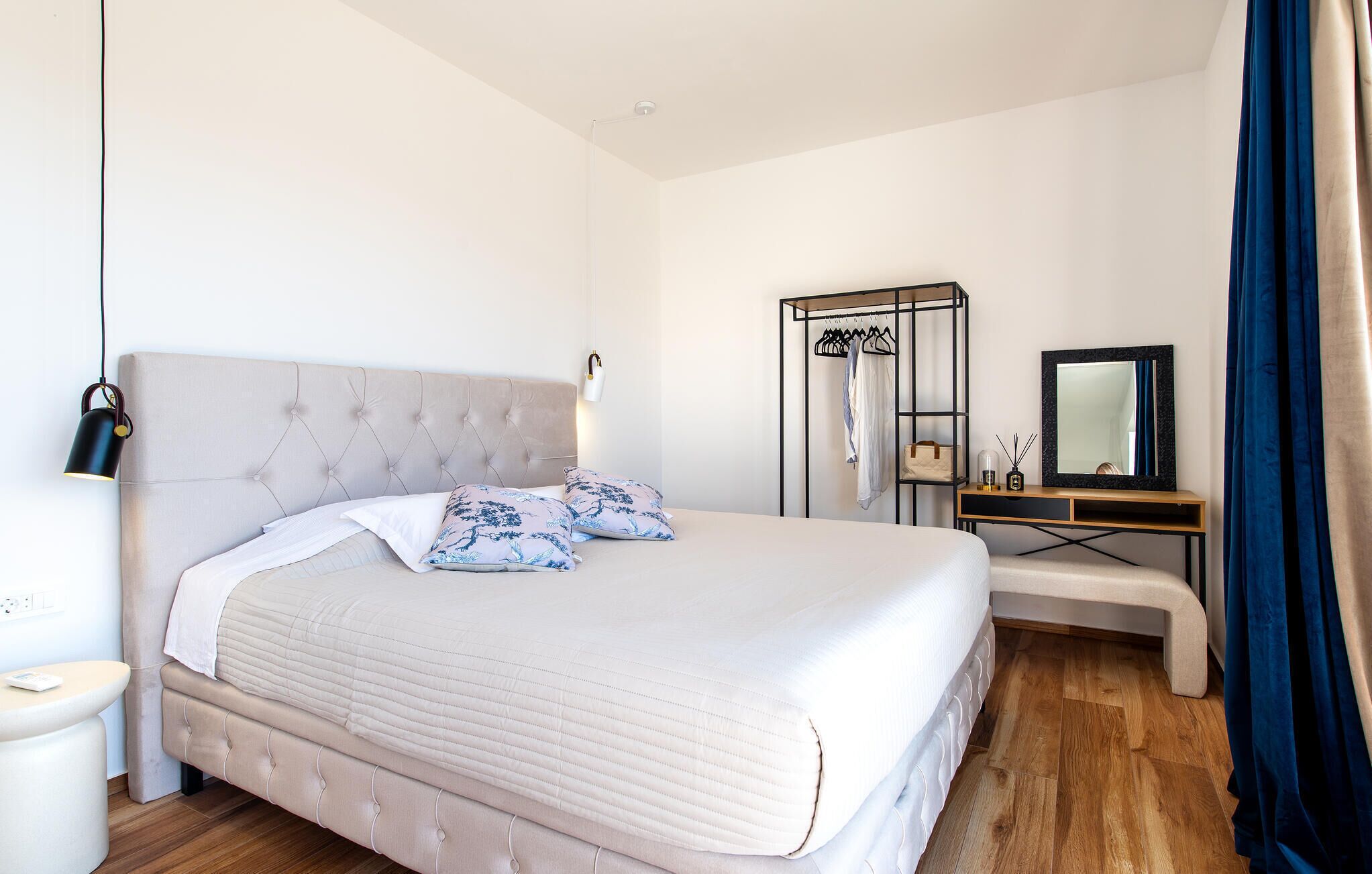 Ein Schlafzimmer mit einem gepolsterten Kopfteil, einem Bett mit weißer Bettwäsche, einem Nachttisch, einer Bank, einem Kleiderschrank, einem Spiegel und einer Pendelleuchte.
