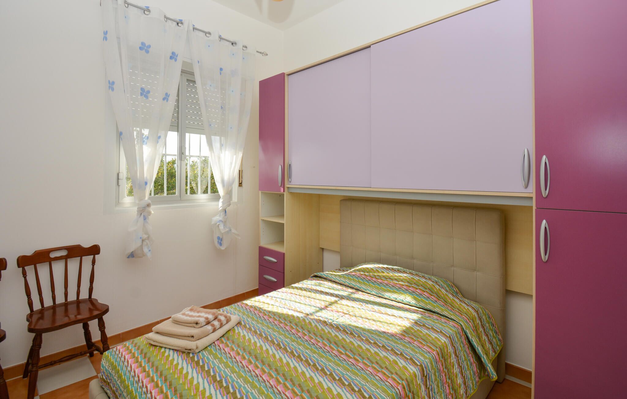 A bedroom with a bed, a chair, a window with curtains, and a wardrobe.