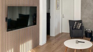 Un saló modern amb un televisor de pantalla plana muntat a una paret de panells de fusta, un sofà negre decoratiu amb un coixí, una taula de café blanca rodona i un petit gaster amb cestes.