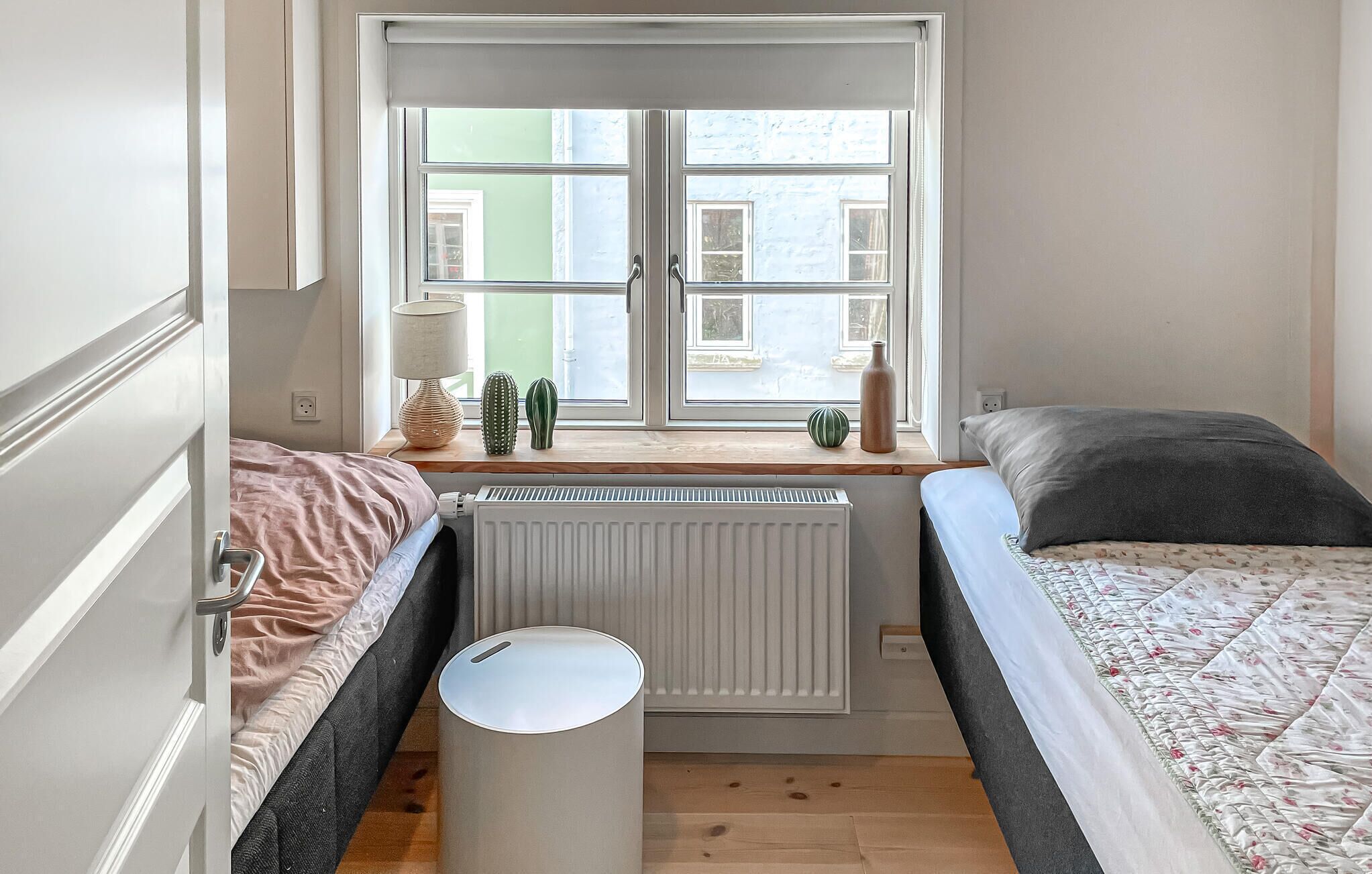 A bedroom with two beds, a radiator, a window with a view of buildings, and a small table with decorative items.