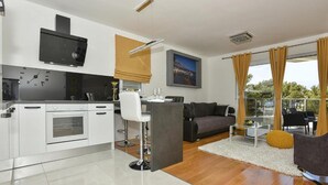 A modern kitchen with white appliances, a dining area with a bar stool, a grey sofa, and a view of the outdoors through large windows. - Nice apartment in Makarska with WiFi (Makarska)