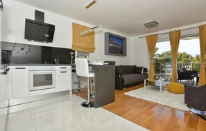 A modern kitchen with white appliances, a dining area with a bar stool, a grey sofa, and a view of the outdoors through large windows. - Nice apartment in Makarska with WiFi (Makarska)