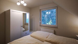 Ein Schlafzimmer mit Bett, Schrank, Fenster mit Blick auf Bäume und einer Deckenleuchte.