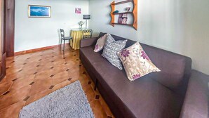 Een woonkamer met een bruine bank, gekleurde kussens, een klein rond tafeltje met een gele tafelkleed en een grijs vloerkleed.