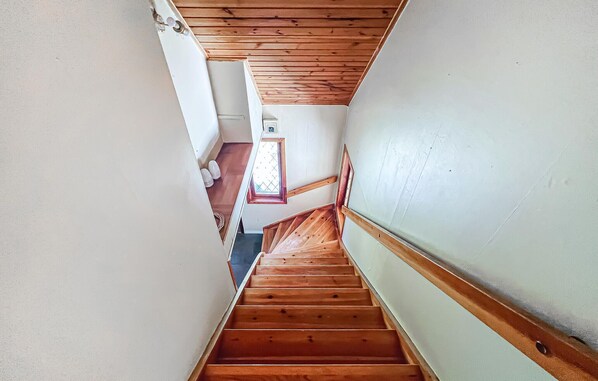 Un escalier en bois, doté d’une rampe, mène à une pièce dont le plafond est également en bois et qui possède une fenêtre à carreaux.