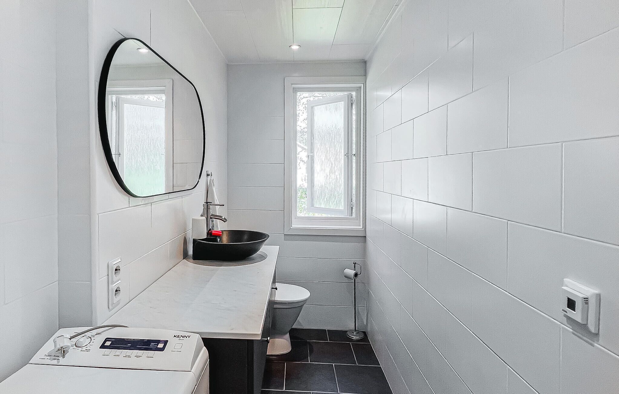 A narrow bathroom with a white sink, a mirror, a toilet, and a window.