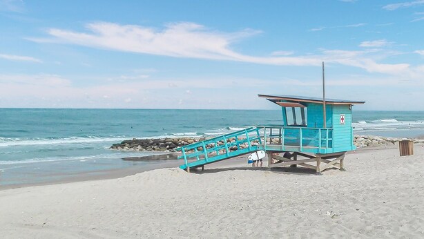 Ein Rettungsschwimmturm mit einer Treppe zu einer Plattform, gelegen an einem Sandstrand mit dem Meer im Hintergrund.