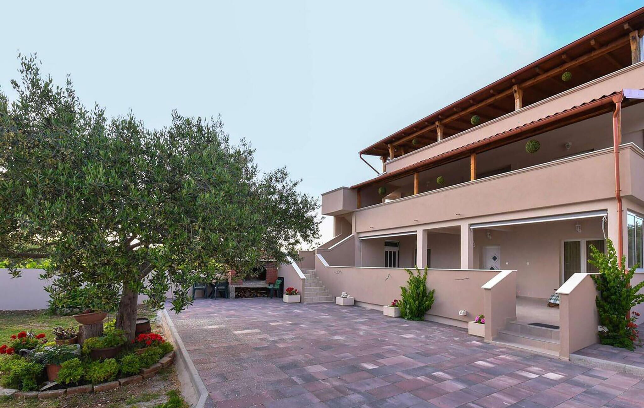 Un edificio de dos pisos con techo plano y un balcón en el nivel superior. Hay un patio empedrado con plantas en macetas y un árbol.