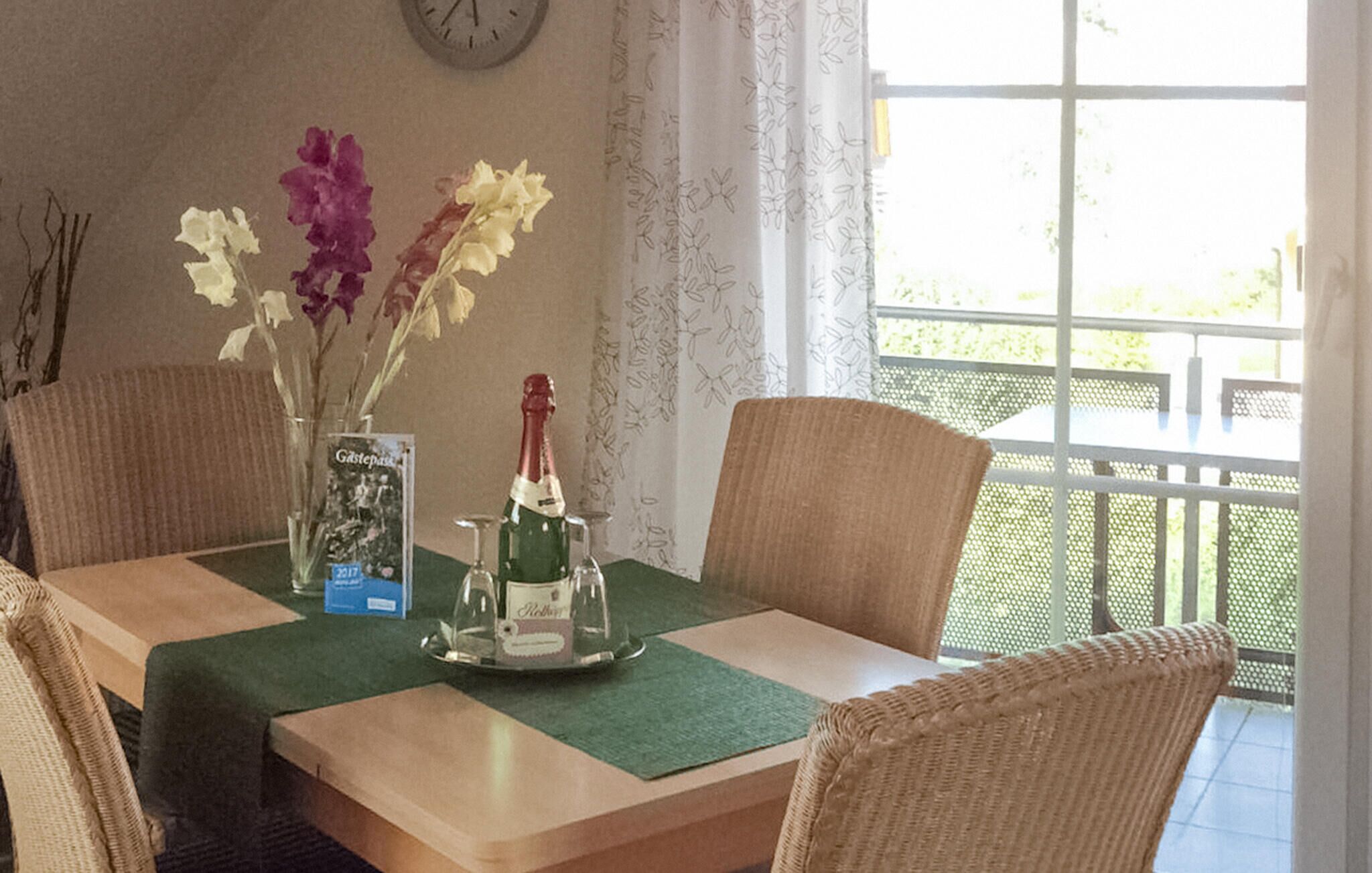 A dining table with a bottle of champagne, a glass, and a menu. There are wicker chairs around the table, and a window with a view of greenery.