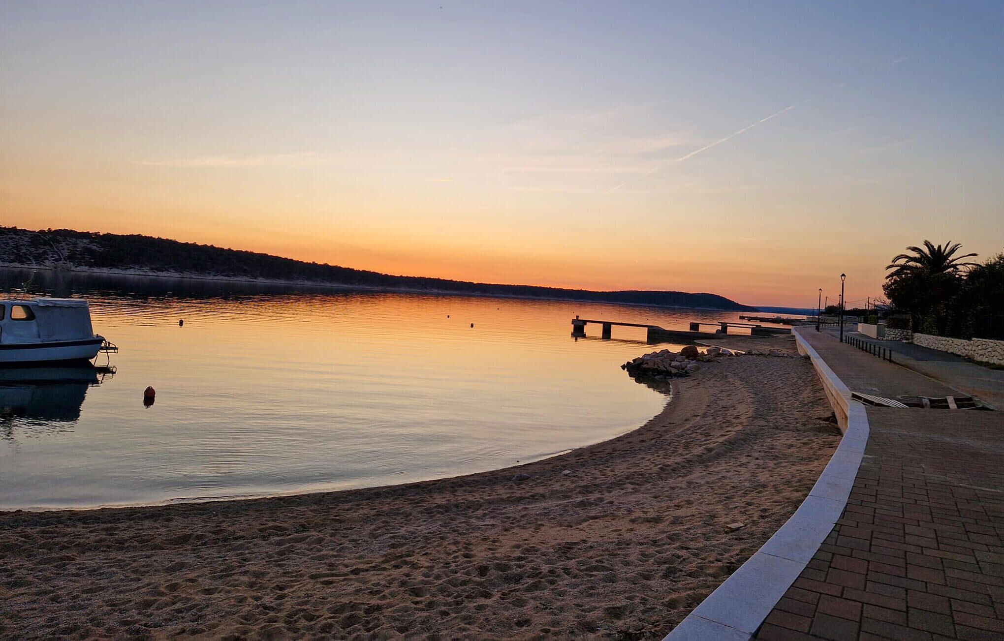 A calm beach with a boat docked on the left, a paved walkway, and a distant shoreline.
