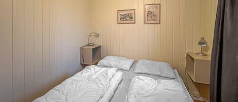Et lille værelse med en enkelt seng, træpanelvæg, to sengeborde med lamper og to billeder i rammer.