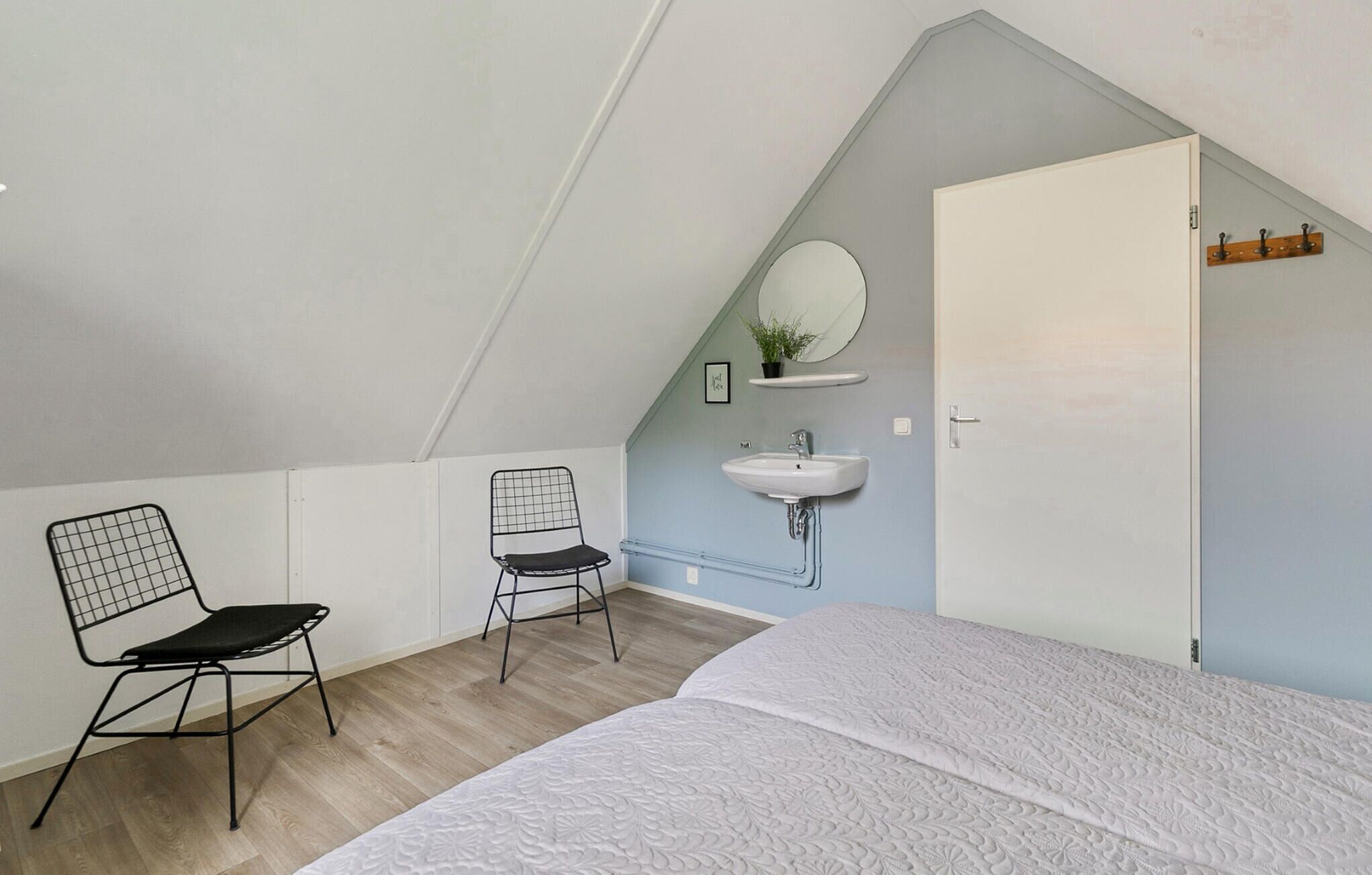 A bedroom with a sloped ceiling, a bed, a chair, a small table, a wall-mounted sink, and a door.
