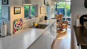 Een moderne keuken met een groot eiland, een spoelbak en een stoel. Er hangen ingelijste foto’s aan de muur en een raam met uitzicht op groen.