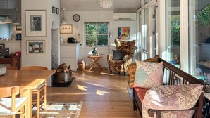 Een knusse woonkamer met houten vloer, een open haard, een eettafel en een groot raam met uitzicht op groen.