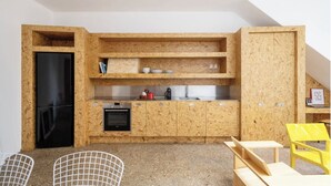 Een compacte keuken met een zwarte koelkast, houten kastjes, een spoelbak en een kleine oven. Er zijn twee stoelen en een gele stoel in de zithoek.