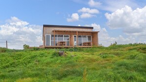 Una cabaña moderna de madera con ventanas grandes y una terraza, rodeada de hierba verde exuberante y flores silvestres.