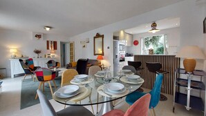 Zona pranzo con tavolo in vetro apparecchiato per sei persone, circondato da sedie colorate. Sullo sfondo, si intravede una cucina con elettrodomestici moderni.