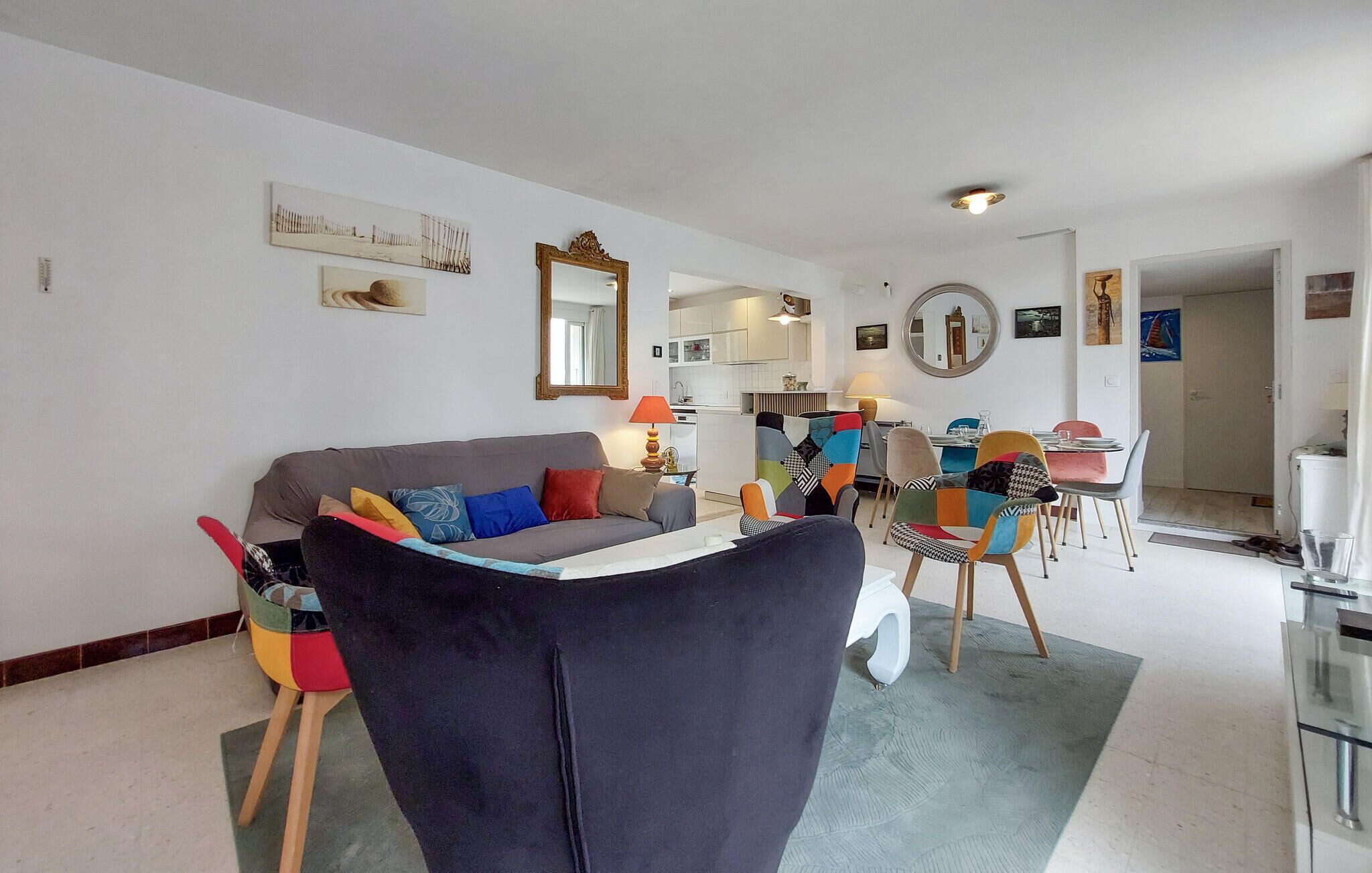 A modern living room with a grey sofa, colorful chairs, a dining table, and a kitchen in the background.