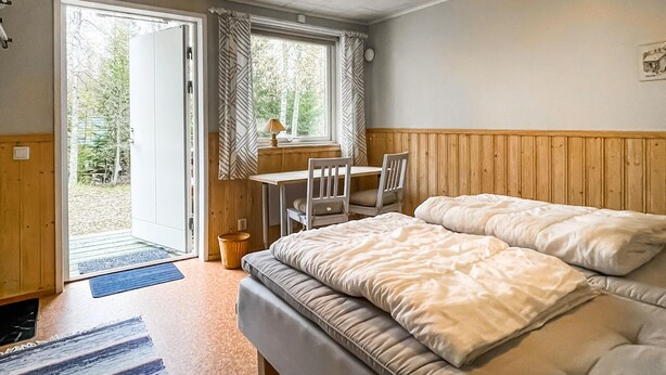 A bedroom with a bed, a desk, and a chair. There is a window with curtains, a door, and a view of greenery outside. - 3 bedroom beautiful home in Torsby (Torsby)