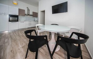 Een moderne keuken met witte kasten, een ingebouwde oven en een roestvrijstalen afzuigkap. Er staat een rond glazen tafel met vier zwarte stoelen en één witte stoel. Een flatscreen televisie is aan de muur boven de tafel bevestigd.