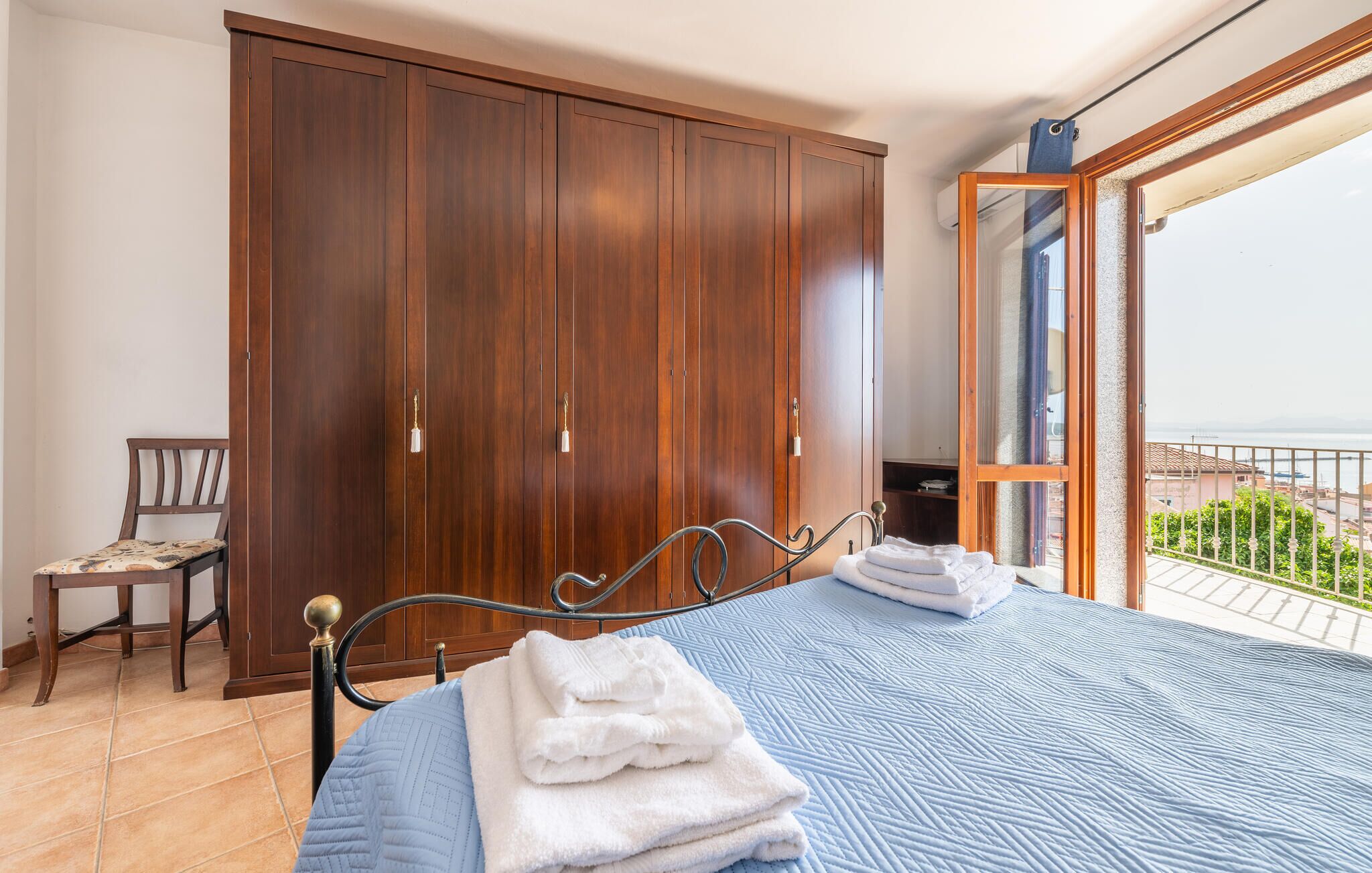 Un dormitorio con un armario grande, una cama con colcha azul y vistas al mar desde un balcón.