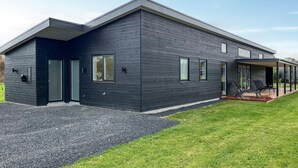 Une maison moderne à deux étages, dotée d’une façade sombre, de grandes fenêtres et d’un espace extérieur aménagé avec un coin salon abrité.