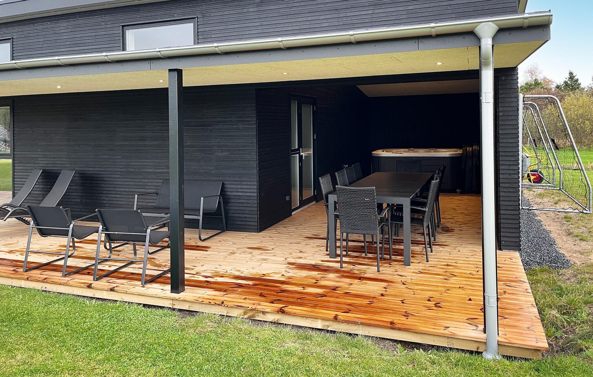 A modern house with a covered patio area featuring a wooden deck, outdoor seating with chairs and a table, and a visible door leading to the interior.