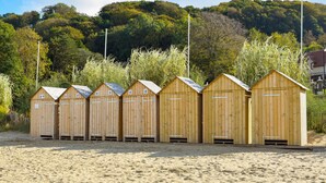 Seis cabañas de madera en la playa, con puertas y ventanas de metal, alineadas sobre una arena con una colina boscosa al fondo.