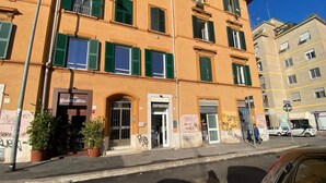 Ein mehrstöckiges Gebäude mit grünen Fensterläden, einem gut sichtbaren Eingang und Pflanzen in Töpfen auf dem Bürgersteig.