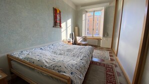 Ein Schlafzimmer mit Bett, einem Sessel, einem Heizkörper und einem Fenster mit Blick auf Gebäude.