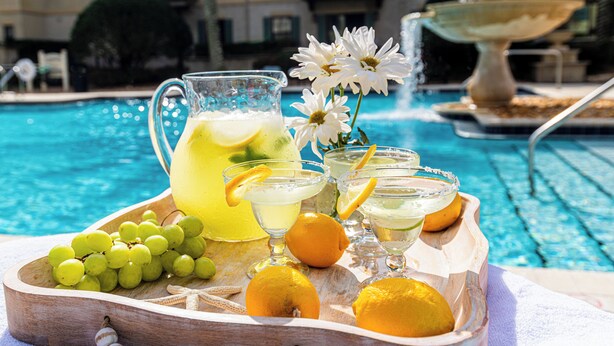 Ett brickbord med en kanna lemonad, glas med drinkar, vindruvor och citroner, placerat vid en pool med en fontän.