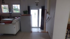 Uma sala de estar ampla, com um sofá branco, uma mesa de centro e um espaço de jantar com uma pequena mesa e cadeiras. Há uma porta de vidro deslizante que dá para uma varanda.