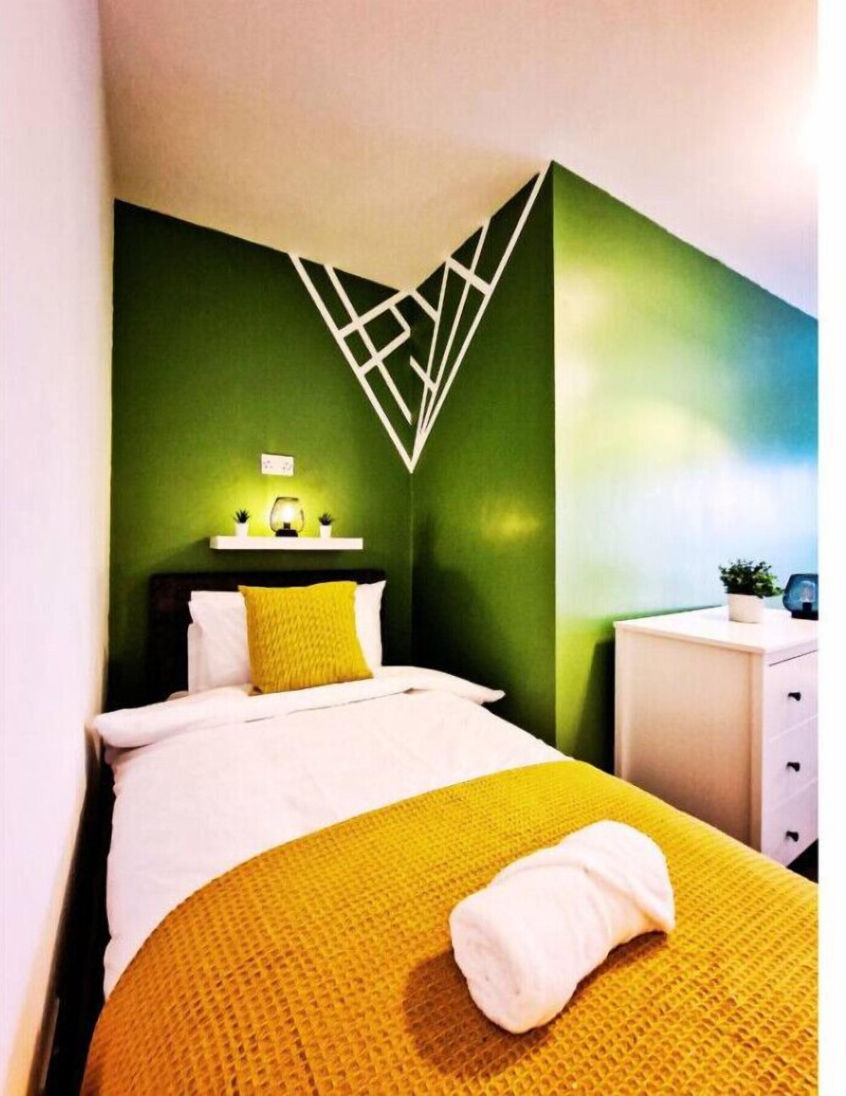 Un lit bien fait, avec une couverture et des oreillers jaunes, une table de chevet blanche avec une lampe, et un motif géométrique sur le mur.