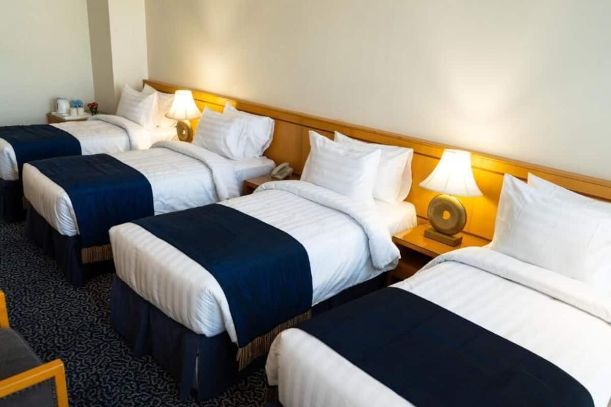 A hotel room with four single beds arranged in a row, each with a bedside table, lamp, and a telephone.