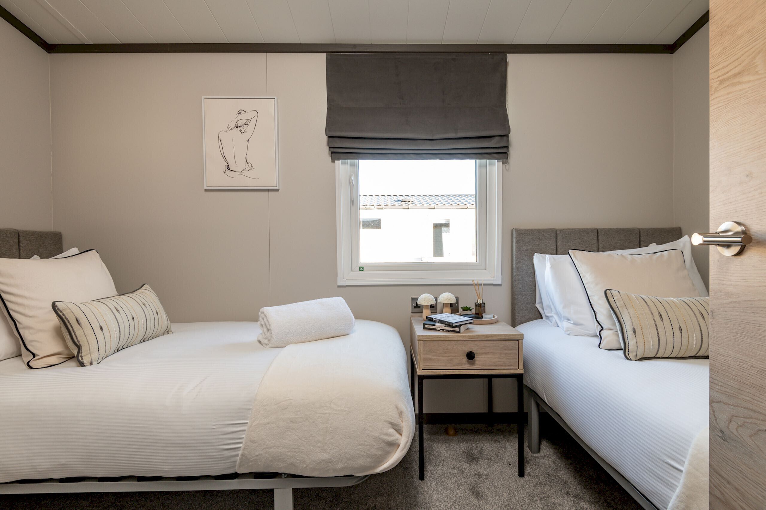 A bedroom with two beds, a window with a view, a bedside table with a lamp, and a framed picture on the wall.
