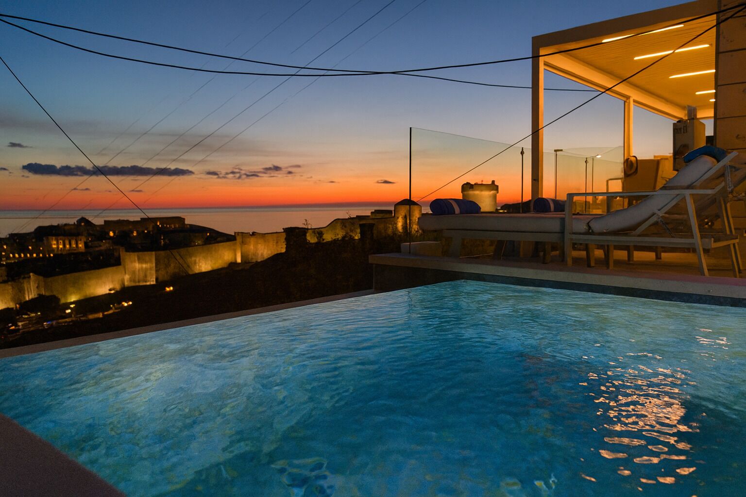 A swimming pool with a sunset view, lounge chairs, and a glass-enclosed area with a view of a historic cityscape.