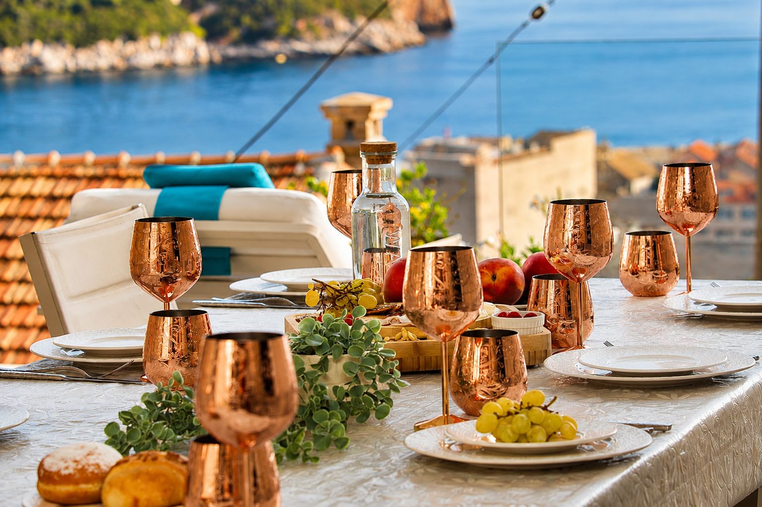 A table set with copper wine glasses, plates, and a bottle, with a scenic coastal view in the background.