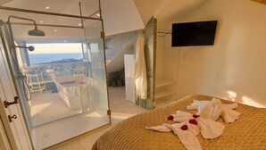 A bedroom with a bed, a glass door leading to a balcony, a TV mounted on the wall, and a view of the sea.