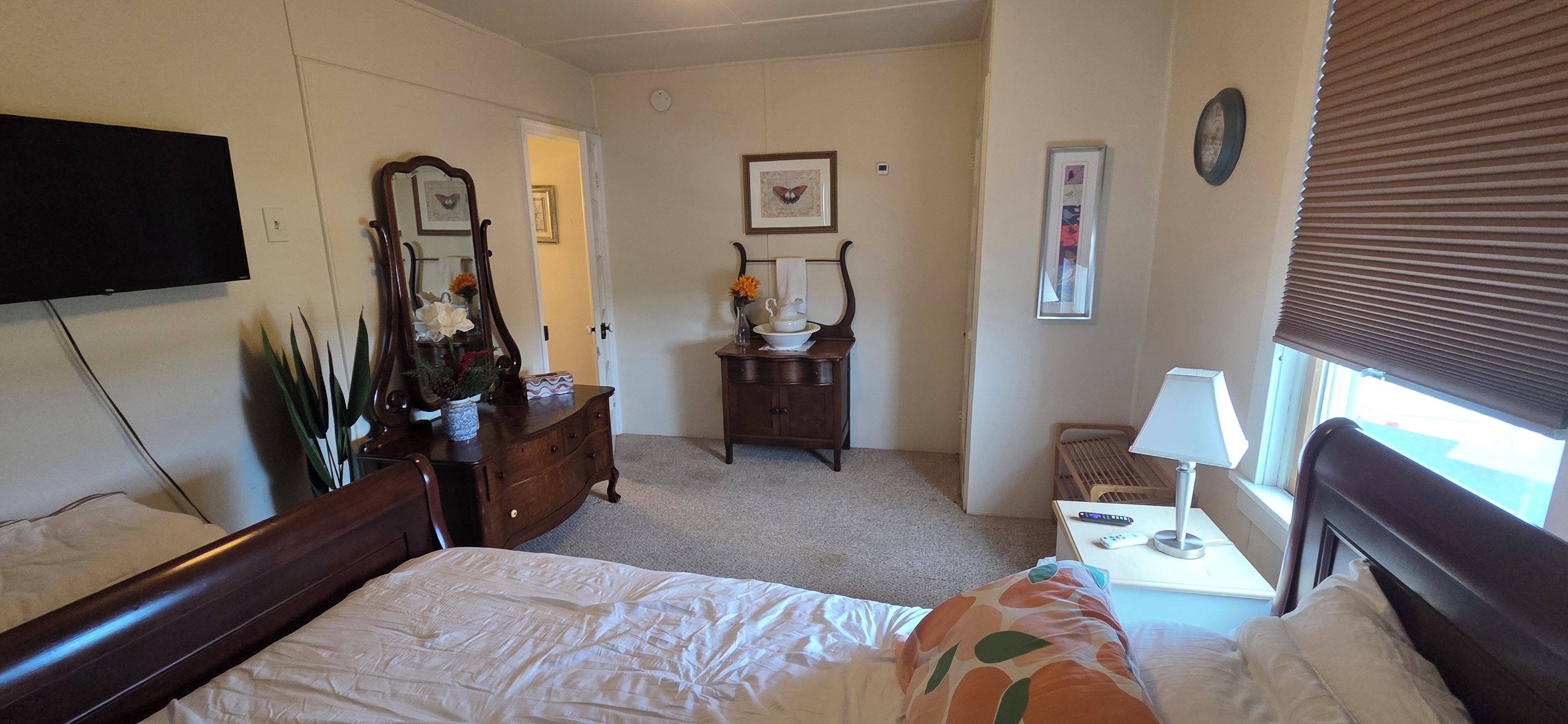 A hotel room with a bed, a dresser, a bedside table with a lamp, a television mounted on the wall, and a window with blinds.