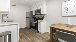 A compact kitchen with white cabinetry, a stainless steel refrigerator, a microwave, an oven, and a sink. There's a dining table with a chair and a modern art piece on the wall. - Landing | Stylish 1BD, Gym (Dunedin)